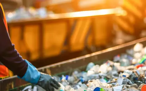 Pracownik w kamizelce odblaskowej sortuje plastikowe butelki na taśmie. Data 191204.