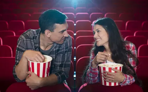 Para w kinie, zajadając popcorn, patrzy na siebie z uśmiechem. Ich spojrzenia sugerują, że to nie tylko film ich interesuje, ale też wzajemna bliskość.