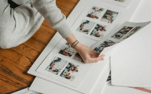 Osoba przegląda wydruki zdjęć, planując, jak zrobić fotoksiążkę. Na papierze widać małe zdjęcia ludzi.