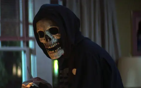 Eine Person mit Totenkopfmaske und Kapuze hält blutige Gegenstände.