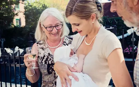 Elegancka mama karmi dziecko na chrzcinach. Babcia z kieliszkiem wina podziwia wnuka. Jak się ubrać na chrzciny? Klasycznie i z klasą.