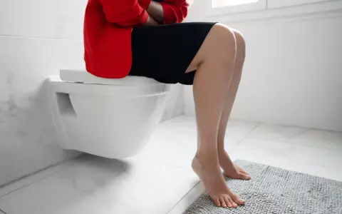 Kobieta w czerwonej marynarce i czarnej sukience siedzi na toalecie, trzymając się za brzuch. Szuka domowych sposobów na skąpomocz.