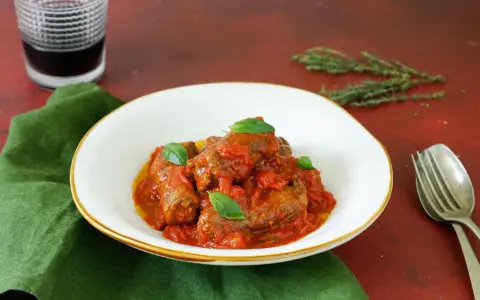 Involtini di carne in padella con sughetto, guarniti con basilico fresco, serviti in un piatto bianco su un tovagliolo verde.