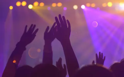 Des mains levées dans la foule d'une boîte de nuit, sous des lumières violettes et jaunes. Le son fort peut causer un acouphène après la boîte de nuit.