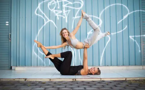 Två kvinnor utför acro yoga framför en blå vägg med graffiti. Den ena ligger på rygg och lyfter den andra som är i en böjd position.