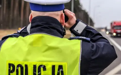 Policjant w kamizelce odblaskowej obserwuje drogę, gdzie widać ciężarówkę. Na drodze obowiązuje zakaz wyprzedzania dla ciężarówek.