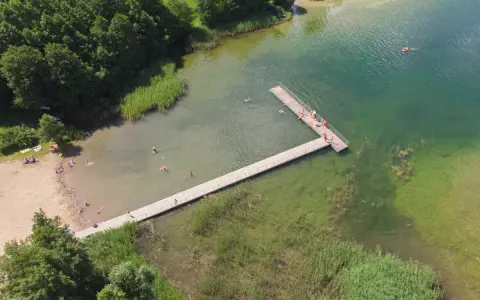 Letnie popołudnie nad jeziorem Hańcza. Ludzie kąpią się i odpoczywają na plaży, inni spacerują po drewnianym pomoście.