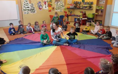 Dzieci i pani bawią się w kole na kolorowym spadochronie. Radosne zabawy w kole dla starszych dzieci w przedszkolu.