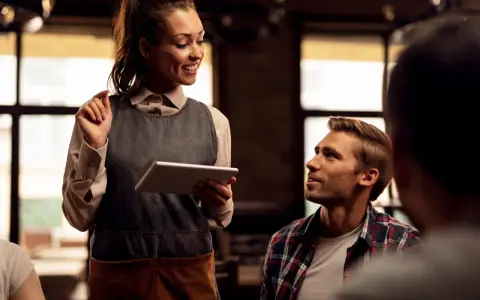 Camarera sonriente atiende a clientes de restaurante, tomando nota en una tablet.