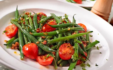 Insalata di fagiolini freschi con pomodorini ciliegino e erbe aromatiche, servita in un piatto bianco.