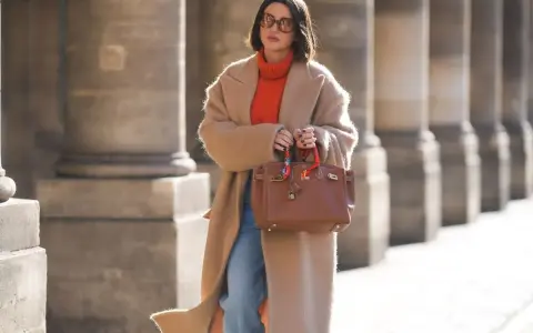 Elegancka kobieta w płaszczu i pomarańczowym golfie, prezentująca modne mom jeans stylizacje zima. Brązowa torebka dodaje szyku.