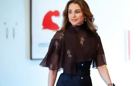 Queen Rania fashion: A woman in a dark brown, floral-patterned top with a high neckline and flared sleeves, paired with a dark skirt.