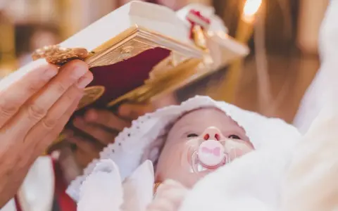 Dziecko z chupkiem w buzi, otulone białym materiałem, podczas ceremonii chrztu. Nad nim trzymana jest księga, a w tle płonie świeca.