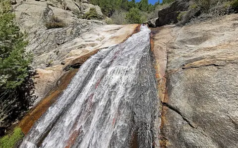 El agua desciende por la roca en la cascada del Hornillo, rodeada de vegetación y rocas graníticas bajo un cielo azul.