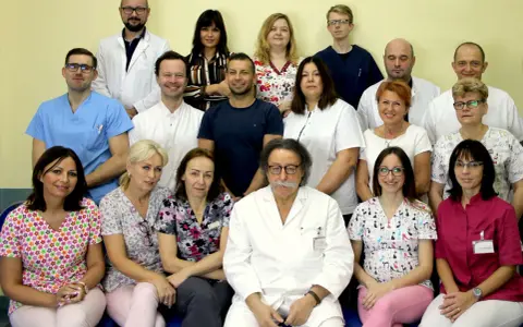 Grupa lekarzy i pielęgniarek w białych fartuchach i kolorowych strojach medycznych, pozująca do zdjęcia.