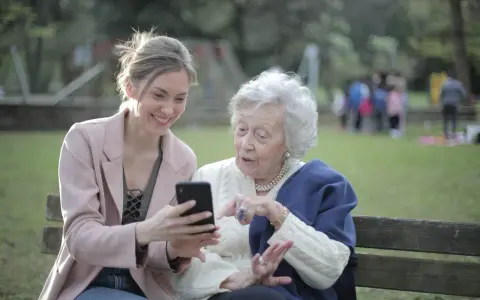 Młoda kobieta pokazuje coś na telefonie starszej pani. To scena z życia, jak z kursu opiekuna osoby starszej, gdzie uczymy się wspierać seniorów w codzienności.