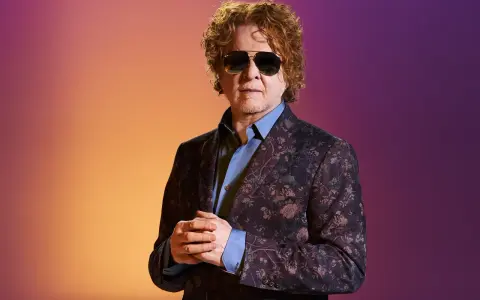 Homme aux cheveux bouclés roux, portant des lunettes de soleil et un costume fleuri, sur fond dégradé orange et violet. Il ressemble à un i monster.