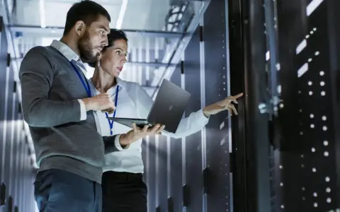 Two IT professionals collaborate on network management, one holding a laptop while the other points to a server rack.