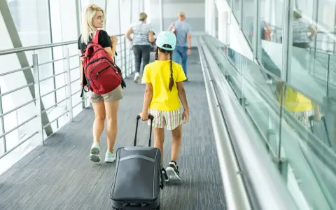 Młoda kobieta z plecakiem i dziewczynka z walizką idą korytarzem lotniska. To może być ich wyjazd nieletniego za granicę bez rodziców.