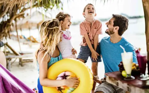 Familie lacht am Strand, genießt die Sommerferien. Ein gelber Schwimmring und Cocktails deuten auf Urlaubsfreude in den Bundesländern hin.
