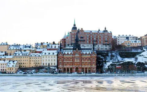 Vinter i Stockholm med snöklädda tak och isig vattenyta. Vackra byggnader reser sig mot en klar himmel.