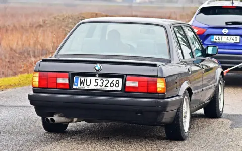 Tył czarnego BMW E30 z tablicą rejestracyjną WU 53268, holowanego przez niebieskiego SUV-a.