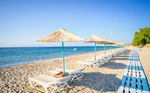 Plaża na wyspie Kos. Rzędy leżaków pod słomianymi parasolami, drewniana ścieżka i spokojne morze.