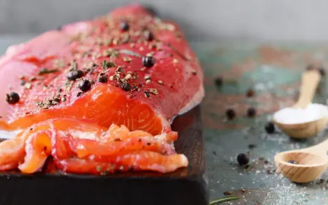 Un filet de gravlax de truite, assaisonné de poivre et d'herbes, prêt à être dégusté. Deux cuillères en bois contiennent sel et genièvre.