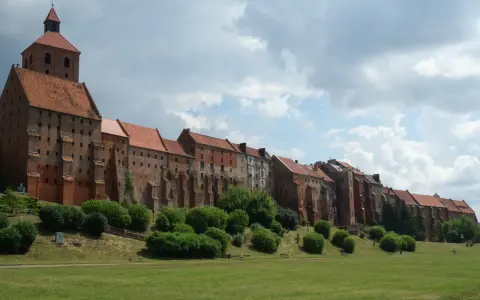 Długi, ceglany zamek w Grudziądzu w jeden dzień. Zielona trawa i krzewy przed zabytkową budowlą.