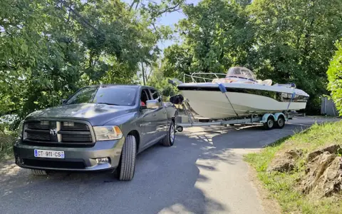 Un pick-up gris est prêt à tracter un bateau de 7m sur sa remorque, sous un ciel bleu et entouré de verdure.