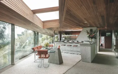 Interno della Sheats Goldstein Residence con soffitto in legno, ampie vetrate, zona pranzo con tavolo in vetro e sedie in pelle arancione, e un bancone bar in cemento con sgabelli.
