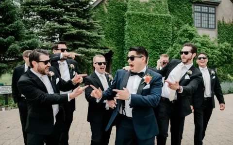 Groomsmen in black suits and cream ties, with the groom in a navy suit, all sporting sunglasses and a playful mood. Do groomsmen match the groom? Not exactly, but they're all dressed to impress!