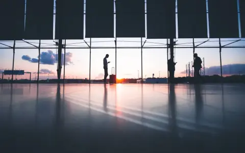 Sylwetki ludzi z bagażami czekają na lotnisku o zachodzie słońca. Czyżby czekał ich overbooking?