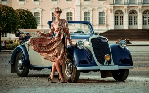 Elegancka kobieta w kwiecistej sukience i okularach przeciwsłonecznych opiera się o zabytkowy samochód. Stylizacja nawiązuje do mody lat 20.