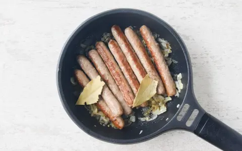 Salchichas doradas cocinándose en sartén con cebolla y hojas de laurel.