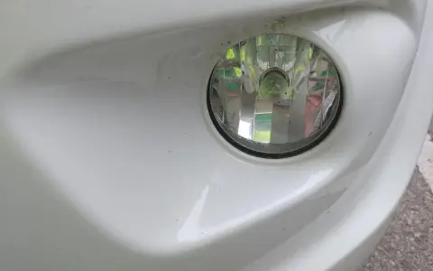 Zbliżenie na biały zderzak samochodu z widocznym okrągłym reflektorem. To światła przeciwmgielne przednie, lekko zakurzone, gotowe do jazdy w trudnych warunkach.