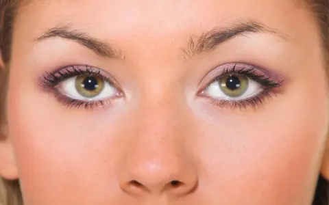 Primo piano di un viso femminile con occhi piccoli e verdi, ciglia lunghe e trucco viola sulle palpebre.
