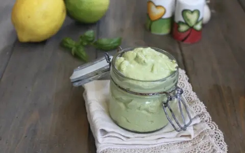 Un vasetto di cremoso pesto al limone, con limoni e basilico sullo sfondo.