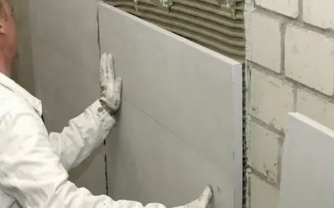 Pracownik w białej kurtce montuje płyty gipsowo-kartonowe, wykonując ocieplanie od wewnątrz. Widać klej na ceglanej ścianie.