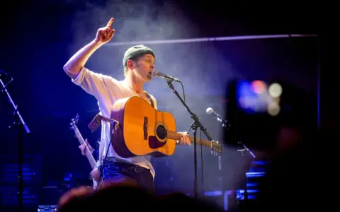 Joel Alme konsert. Sångaren med gitarr och mössa pekar uppåt under en blåbelyst spelning.