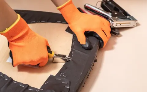 Manos con guantes naranjas usando una grapadora para tapizar una silla.