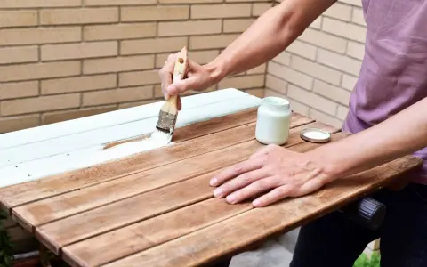Persona pinta madera de blanco con brocha. Un frasco de pintura blanca y su tapa están cerca.