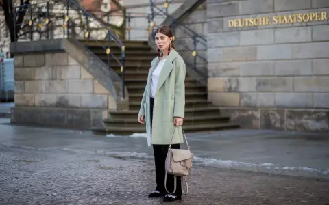 Kobieta w płaszczu w pistacjowym kolorze, z beżową torebką i ozdobnymi butami, stoi przed budynkiem Deutsche Staatsoper.