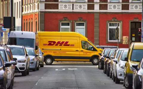 Żółta furgonetka DHL stoi zaparkowana na ulicy, obok innych samochodów. To miejsce, do którego pracują kurierzy DHL, by dostarczać paczki.