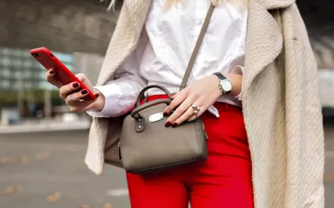 Une femme élégante, avec un manteau beige, un chemisier blanc et un pantalon rouge vif, tient un téléphone rouge et un petit sac gris. Quel entoilage pour sac choisir pour un tel look ?