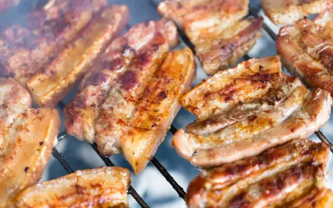 Kawałki soczystego boczku surowego pieką się na grillu, otoczone dymem.