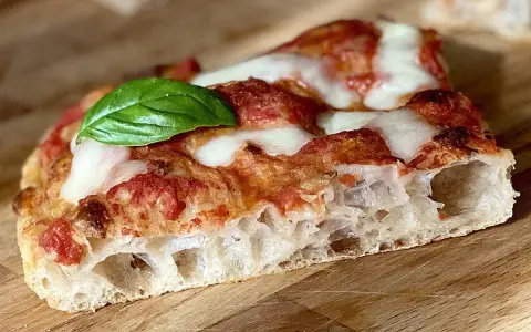 Fetta di pizza fatta in casa teglia, con crosta alveolata, pomodoro, mozzarella filante e una foglia di basilico fresco.