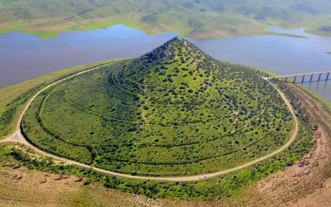 El **cerro Masatrigo**, cubierto de vegetación, se alza majestuoso junto a un embalse. Una carretera serpentea a su alrededor, conectando con un puente.