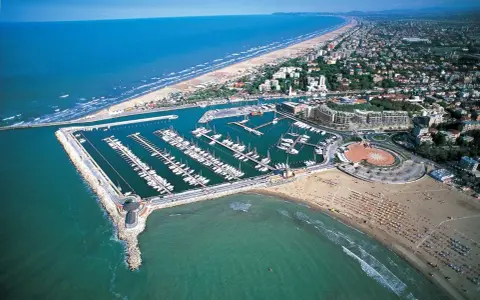 La darsena di Rimini, con le sue barche ormeggiate, si estende lungo la costa adriatica, tra la spiaggia affollata e il mare azzurro.