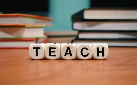 Kolejność wprowadzania liter w przedszkolu mac. Kostki z napisem TEACH na drewnianym blacie, w tle stosy książek.